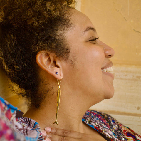 Woman with a smile on her face against a beige wall wearing a long slender gold toned fish earring