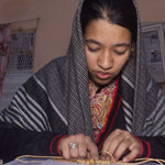 Load image into Gallery viewer, A woman wearing a black and white patterned shawl and a red paisley top sits indoors, working on an ornament with gold-colored thread and beads.
