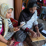 Load image into Gallery viewer, Two women sitting indoors, one holding a phone and the other wrapping or tying a piece of string or thread. The setting is a home interior with furniture and textiles visible in the background.
