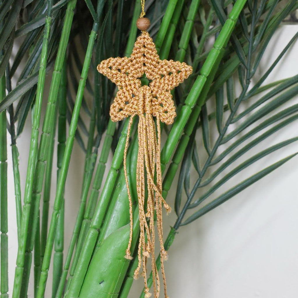 A handmade tan and gold macrame star ornament with a wooden bead top and long dangling tassels, hanging against a background of green faux bamboo stalks.