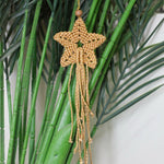 Load image into Gallery viewer, A handmade tan and gold macrame star ornament with a wooden bead top and long dangling tassels, hanging against a background of green faux bamboo stalks.
