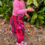 Load image into Gallery viewer, Child wearing a pink sweater and pink, red and black patterned pants standing outdoors with green foliage in the background.
