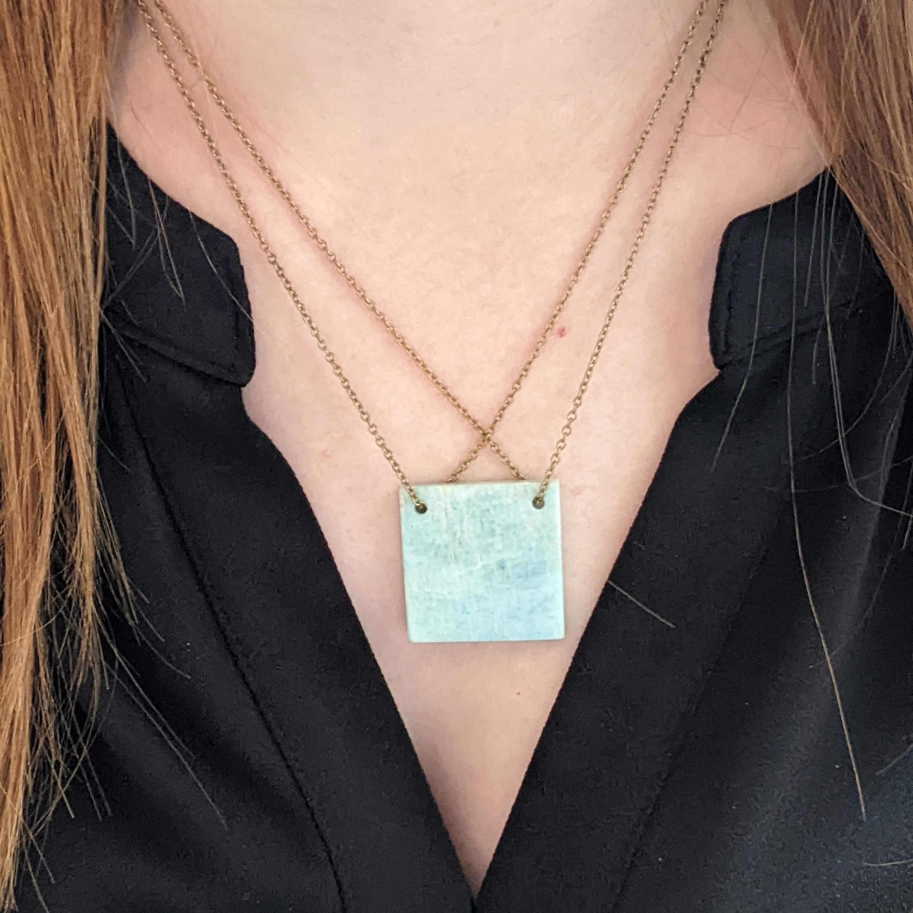 Person wearing a necklace with a turquoise colored square pendant against a black background