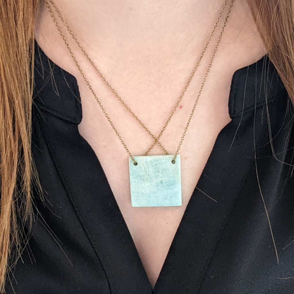 Person wearing a necklace with a turquoise colored square pendant against a black background