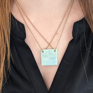 Person wearing a necklace with a turquoise colored square pendant against a black background